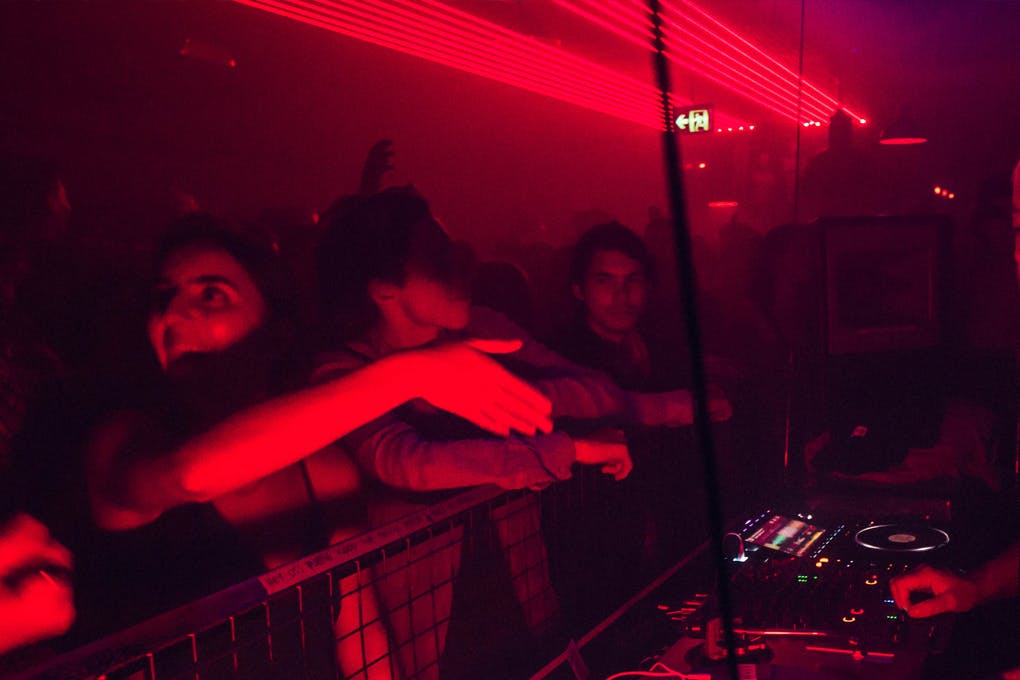 A red-lit nightclub with people dancing and DJ behind the decks.